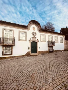 Edificação histórica de fachada branca com detalhes em pedra, porta central verde, janelas simétricas e varandas de ferro forjado. Rua de paralelepípedos ocupa o primeiro plano sob céu parcialmente nublado;