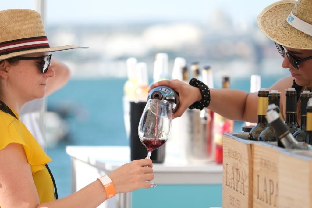 Dois homens de chapéu de palha e óculos escuros, um serve vinho tinto em taça, garrafas em caixas de madeira ao lado.
