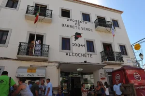 Fachada de prédio com varandas, bandeiras e inscrição “Aposento do Barrete Verde 20-8-44 Alcochete”, pessoas reunidas em frente ao café Nicola.