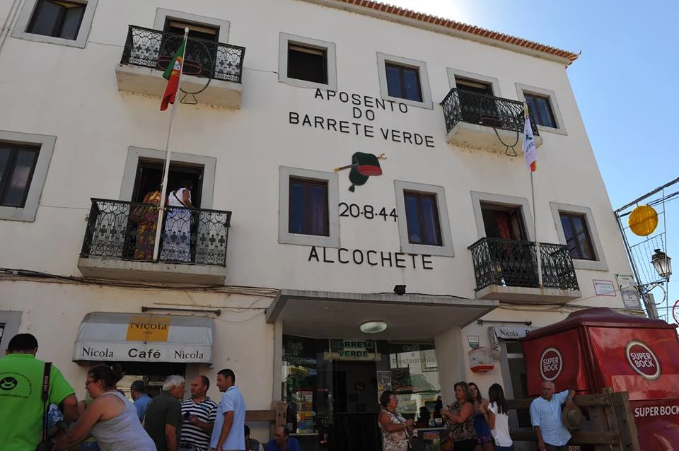 Fachada de prédio com varandas, bandeiras e inscrição “Aposento do Barrete Verde 20-8-44 Alcochete”, pessoas reunidas em frente ao café Nicola.