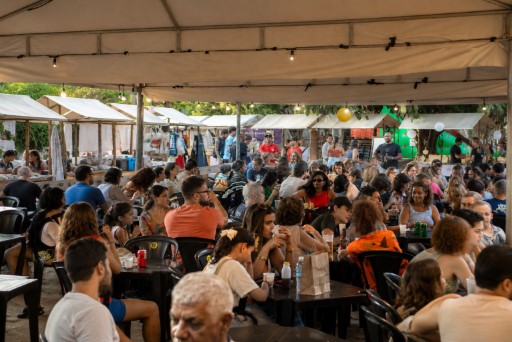 Pessoas sentadas em mesas de madeira sob bandeirolas coloridas e luzes, com pratos e bebidas, em ambiente festivo cercado por árvores e prédios.