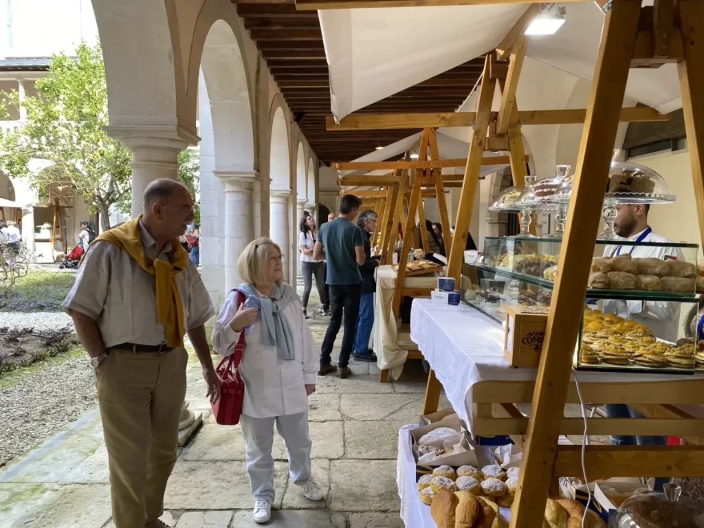 Pessoas caminham por feira coberta em pátio com arcos de pedra, observando barracas de pães e doces expostos em vitrines sobre mesas brancas.