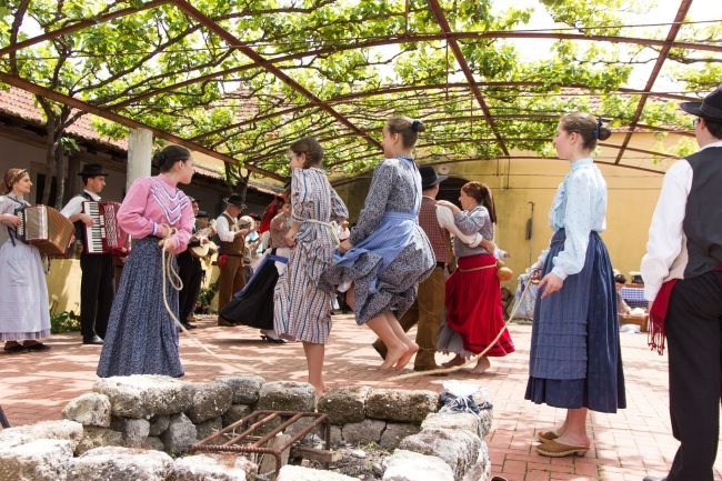 Grupo em trajes regionais dança em pátio de tijolos, músicos com acordeões ao fundo sob pergolado coberto de vinhas.