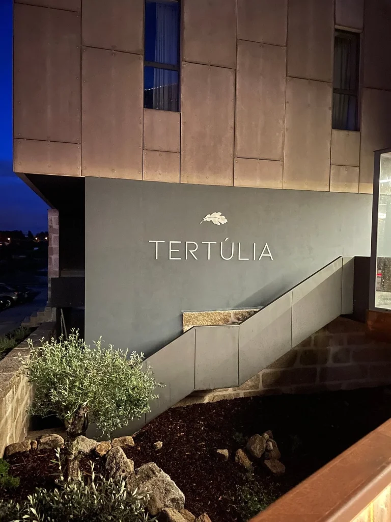 Fachada moderna do restaurante Tertúlia com revestimento metálico, rampa de acesso, jardim com pedras e iluminação noturna. Texto visível: “TERTÚLIA”.