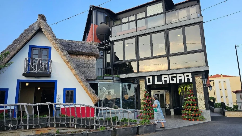 Fachada do restaurante O Lagar com prédio envidraçado de vários andares ao lado de construção com telhado de colmo. Pessoa caminha em frente à entrada decorada com vasos de flores;