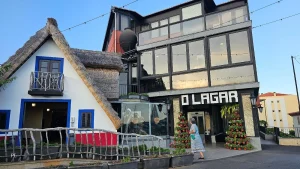 Fachada do restaurante O Lagar com prédio envidraçado de vários andares ao lado de construção com telhado de colmo. Pessoa caminha em frente à entrada decorada com vasos de flores;