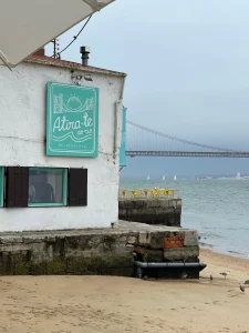 Prédio branco com janelas azul-turquesa e placa “Atira-te ao rio Restaurante”, situado na areia da praia com ponte suspensa e veleiros ao fundo.