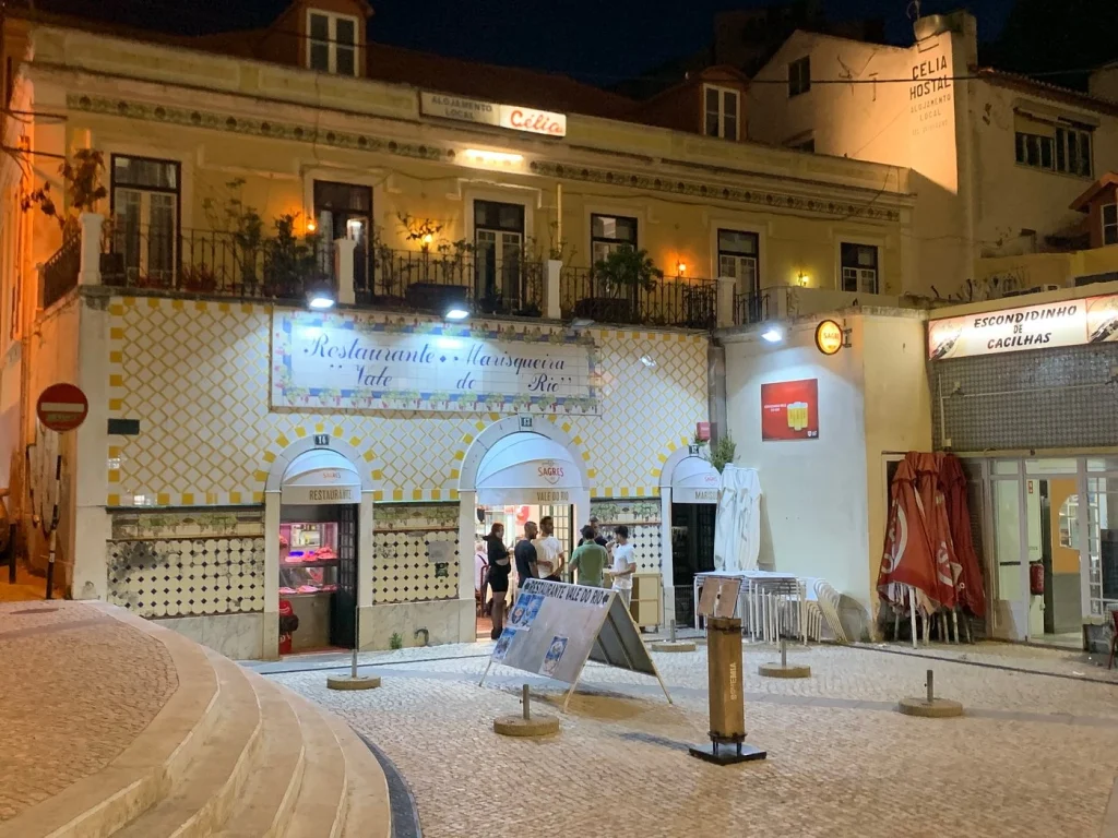 Fachada com azulejos amarelos e brancos, entradas em arco, placa “Restaurante Marisqueira Vale do Rio”, pessoas na porta e luzes quentes.