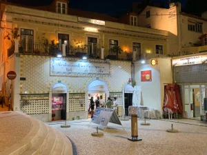 Fachada com azulejos amarelos e brancos, entradas em arco, placa “Restaurante Marisqueira Vale do Rio”, pessoas na porta e luzes quentes.