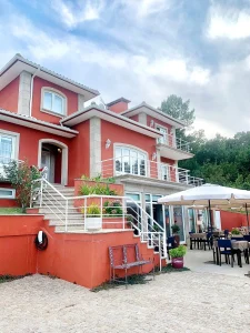 Residência de dois andares em vermelho terracota com varandas brancas, escadaria decorada com plantas e área externa com mesas sob guarda-sol.