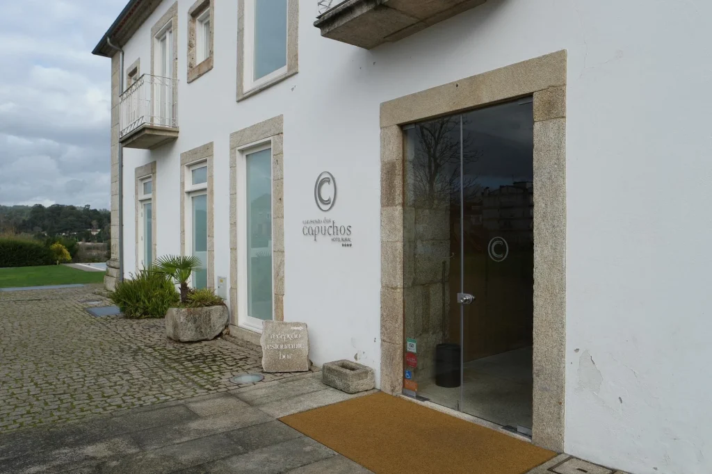Fachada branca moderna com molduras em pedra, porta de vidro, placa “Vila Galé Collection Capuchos Hotel, Nazaré” e bloco “receção restaurante bar”.