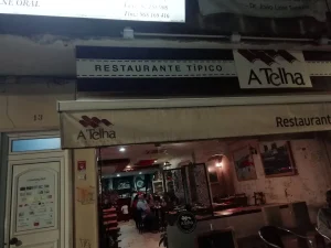 Entrada com toldo e placa “A Telha Restaurante Típico”, número 13 visível, interior iluminado com mesas ocupadas e bandeira portuguesa pendurada.
