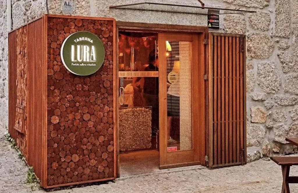 Entrada com parede coberta por rodelas de madeira, porta e moldura em tom natural, placa “Taberna Lura” com subtítulo “Produtos naturais e tradicionais