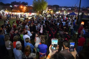 Multidão em círculo assiste grupo de músicos em preto e vermelho tocando tambores à noite, com barracas iluminadas ao fundo.