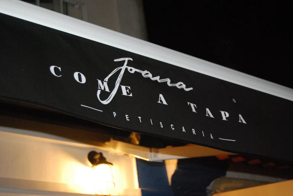 Toldo preto iluminado com texto branco “Joana Come a Tapa Petiscaria”, luz de parede abaixo destacando a entrada do restaurante à noite.
