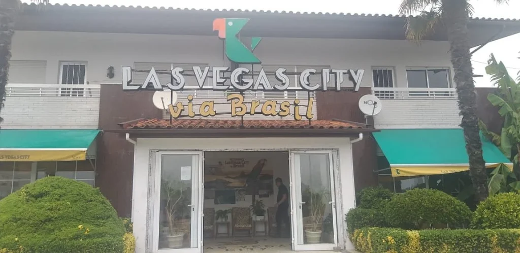 Fachada de restaurante com entrada central envidraçada, jardins laterais e letreiro com os textos “LAS VEGAS CITY” e “Via Brasil”.