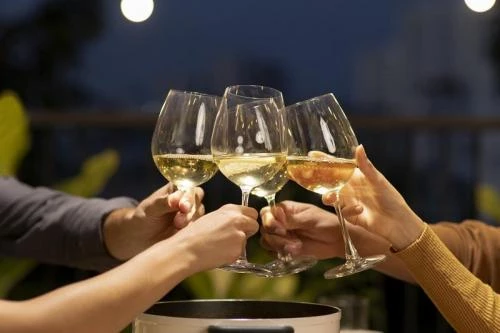 Quatro mãos erguem taças de vinho branco em brinde ao ar livre, com luzes desfocadas e vegetação ao fundo, sugerindo celebração noturna.