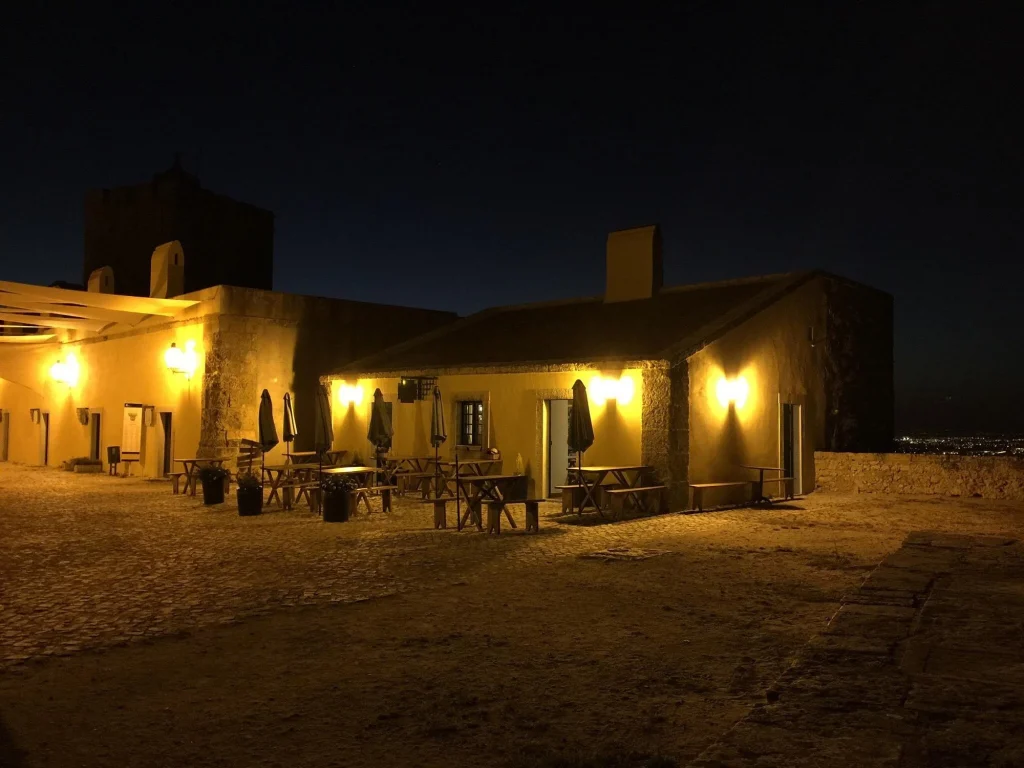 Edificação de estilo histórico iluminada por arandelas amarelas, com mesas e bancos em pátio de pedra sob céu noturno escuro;