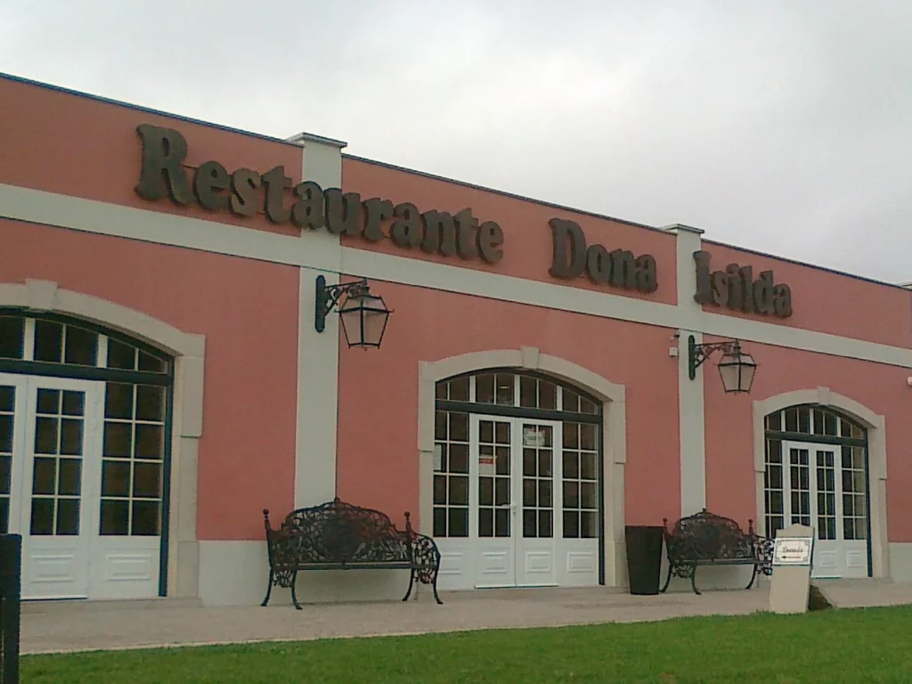 Fachada em tom rosado com o letreiro “Restaurante Dona Isilda”, portas envidraçadas, bancos ornamentais e gramado frontal sob céu nublado;