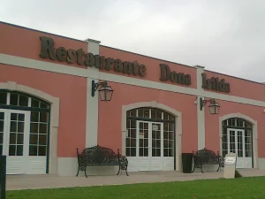 Fachada em tom rosado com o letreiro “Restaurante Dona Isilda”, portas envidraçadas, bancos ornamentais e gramado frontal sob céu nublado;