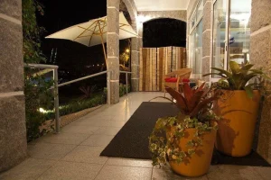 Pátio com piso de azulejos, vasos amarelos com plantas, cadeira de vime com almofada vermelha e guarda-sol branco sob luz quente.