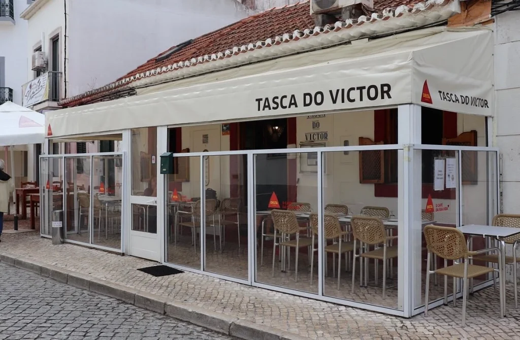 Restaurante com toldo creme escrito “Tasca do Victor”, logotipo Delta Café, área envidraçada com mesas e pessoa passando na calçada.