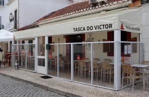 Restaurante com toldo creme escrito “Tasca do Victor”, logotipo Delta Café, área envidraçada com mesas e pessoa passando na calçada.