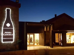 Fachada de edifício iluminado ao anoitecer, com letreiro em formato de garrafa exibindo o texto “PEDRA CANCELA”. Entrada envidraçada e loja de vinhos iluminada aparecem à direita;