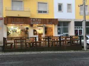 Fachada em madeira com nome “Pátio da Serra”, áreas externas com mesas e bancos, prédio amarelo ao fundo e rua de paralelepípedos com carro estacionado.