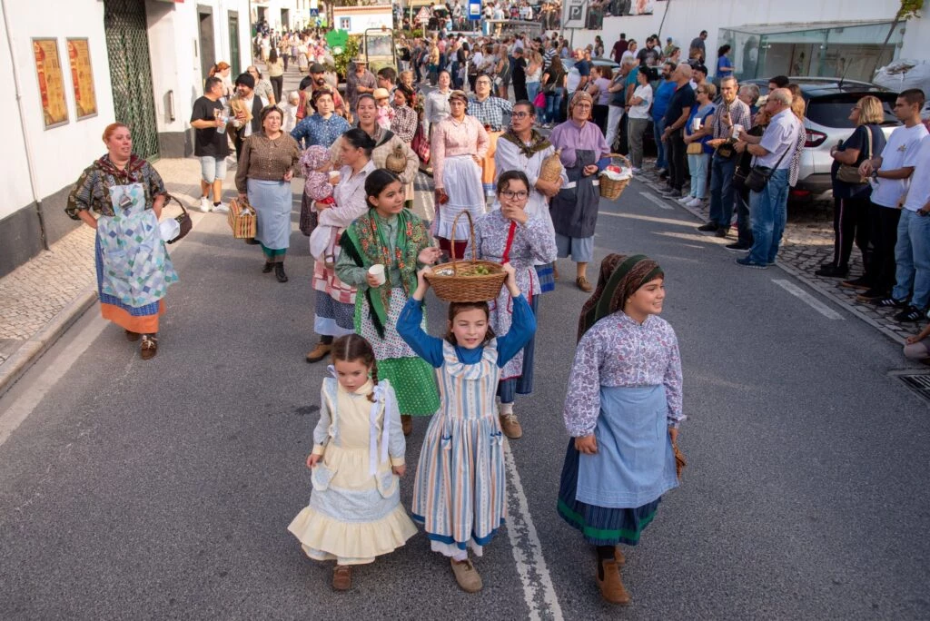 Grupo de pessoas, incluindo crianças, participa de desfile em rua urbana usando trajes tradicionais e carregando cestos, observado por público nas calçadas.