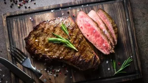Bife grelhado com crosta dourada, decorado com alecrim, acompanhado de fatias com interior rosado sobre uma tábua de madeira.