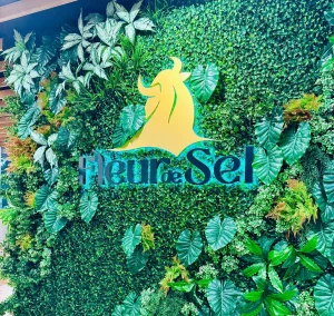 Painel revestido por folhagens ornamentais exibe logotipo dourado e o nome “Fleur de Sel”, compondo decoração interna de ambiente gastronômico.
