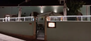 achada de restaurante iluminada à noite, com esplanada elevada, guarda-sóis fechados, mesas ao fundo e acesso por escada central.