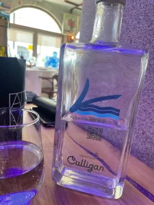 Garrafa de vidro transparente com logotipo "Varanda do Mondego" e QR code sobre mesa de restaurante, ao lado de copo; salão iluminado ao fundo.