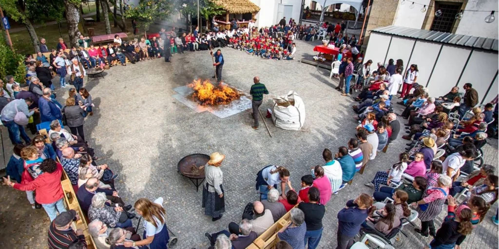 Praça com grande público reunido em círculo ao redor de uma fogueira, acompanhando atividade tradicional ao ar livre durante festival.
