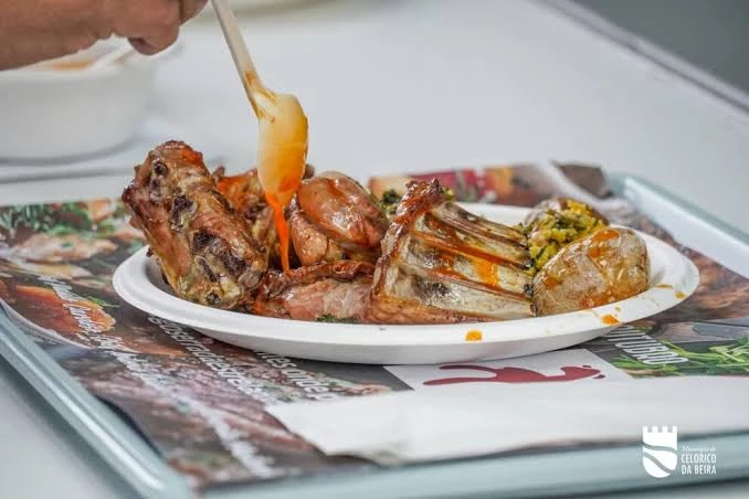 Prato com peças de carne de borrego assada e batatas, regadas com molho alaranjado servido por uma pessoa em bandeja de refeição.