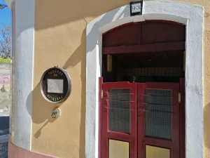 Fachada da Taberna do Quinzena com porta vermelha de duas folhas, moldura branca e quadro circular exibindo o nome e o menu do restaurante.