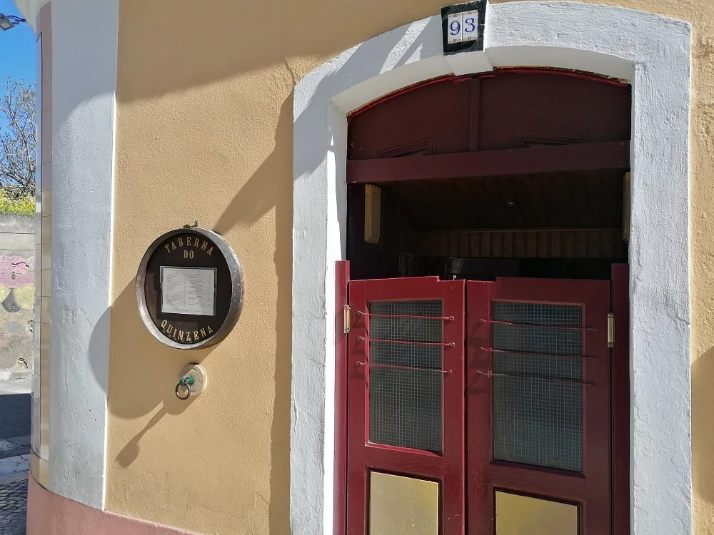 Fachada da Taberna do Quinzena com porta vermelha de duas folhas, moldura branca e quadro circular exibindo o nome e o menu do restaurante.