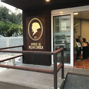 Entrada do restaurante Amigos da Velha Caroca com rampa de acesso, porta de vidro e painel iluminado com a inscrição “1993 Amigos da Velha Caroca”.
