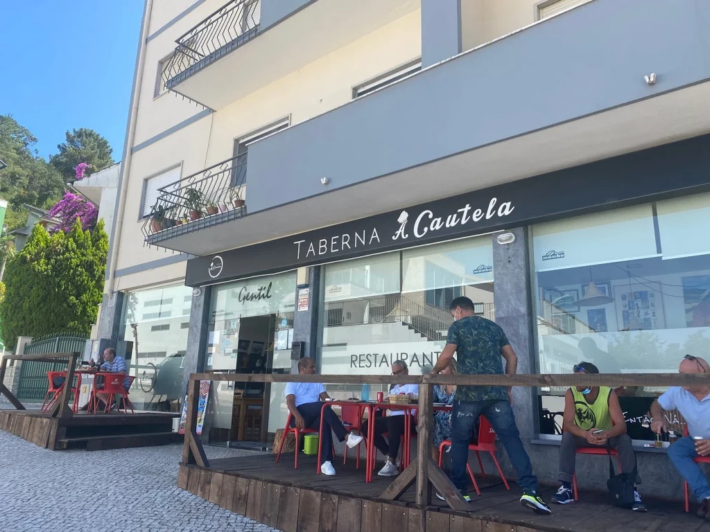 Fachada da Taberna À Cautela com mesas externas ocupadas por clientes, letreiro do restaurante, ampla vitrine e prédio residencial ao fundo.