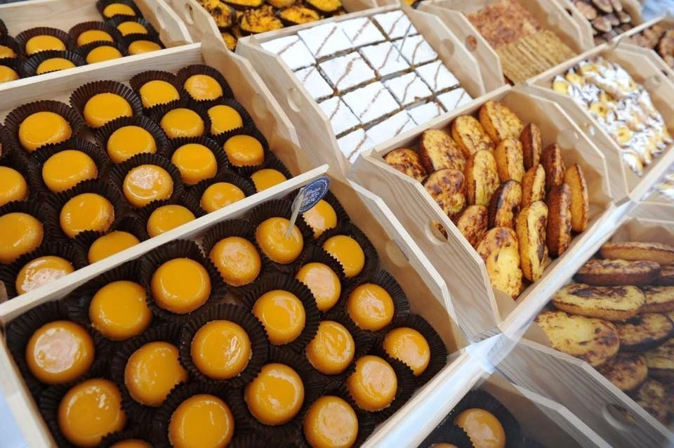 Bandejas de madeira exibem doces e biscoitos variados, com destaque para pastéis de nata organizados em fileiras sobre balcão de exposição.