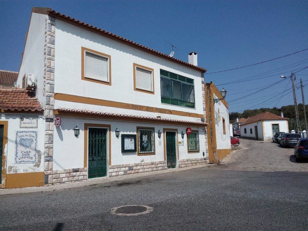 Edifício de dois andares com fachada branca e detalhes ocres, identificado como Casa dos Vinhos, localizado em esquina de rua pavimentada.