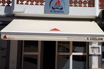 A imagem mostra a fachada do Restaurante A Grelha, em Arganil. O edifício apresenta paredes claras, um toldo com a marca Delta e o nome do restaurante, além do logótipo com um peixe estilizado. Em frente à entrada existe uma mesa em formato de barril e uma cadeira verde.
