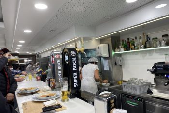 Balcão de restaurante com clientes sentados e em pé, com torneiras de cerveja, pratos, copos e funcionário ao fundo.