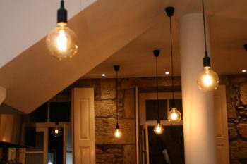 Sala com lâmpadas pendentes de luz quente, teto branco com coluna central, balcão com copos, pratos e desinfetante; ao fundo paredes de pedra, portas abertas e mais iluminação.