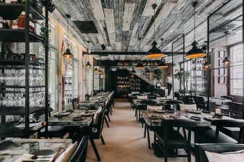 Sala de restaurante com mesas de madeira, cadeiras pretas, iluminação suspensa e divisórias metálicas.