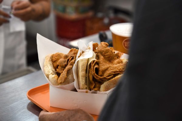 Duas sandes de carne frita em pão branco, colocadas num tabuleiro laranja com um copo de bebida ao fundo.