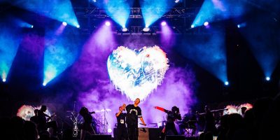 Palco com luzes coloridas e ecrã em forma de coração, com artistas a atuar perante o público.