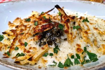 Travessa branca com prato gratinado de aspeto cremoso, com queijo dourado e ervas picadas por cima.
