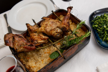 Travessa com carne assada colocada sobre arroz com ervas, acompanhada por um prato de legumes verdes e dois copos de vinho tinto.